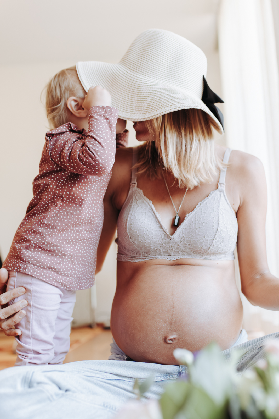 Werdende Mama mit nacktem Babybauch und Hut gibt ihrer älteren Tochter ein Küssen.