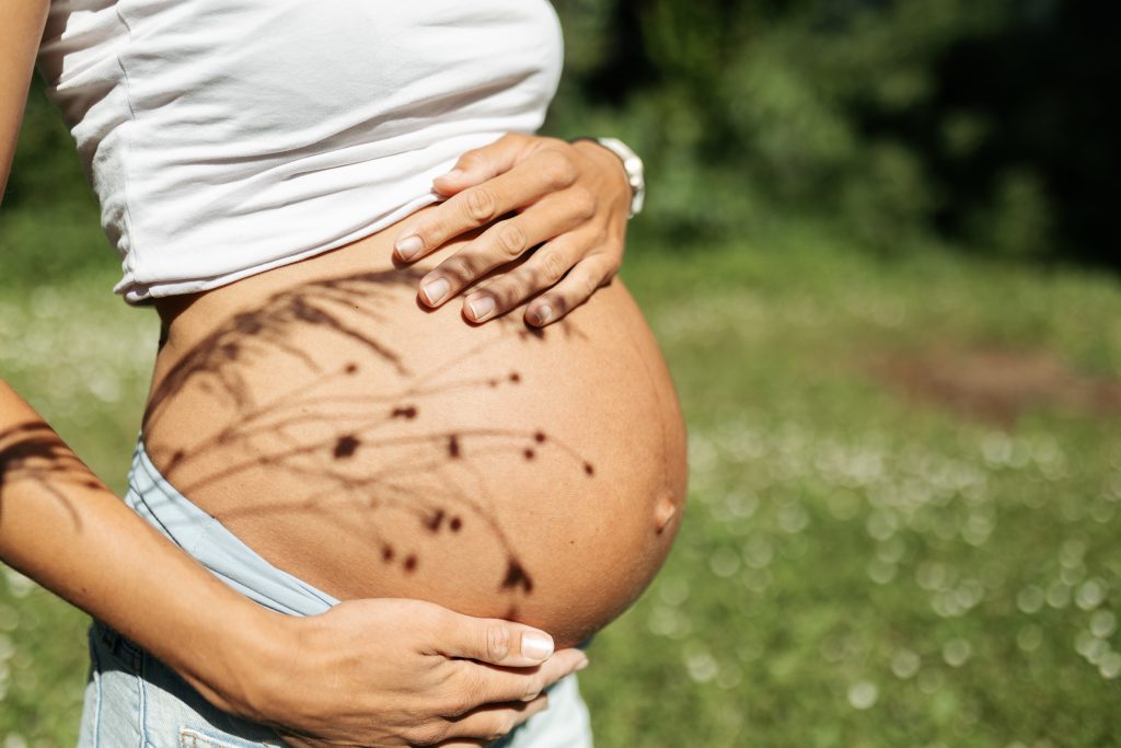 Nahaufnahme eines Babybauches im eigenen Garten im Kaiserstuhl. Auf dem Bauch sind Schatten von Gräsern zu sehen.