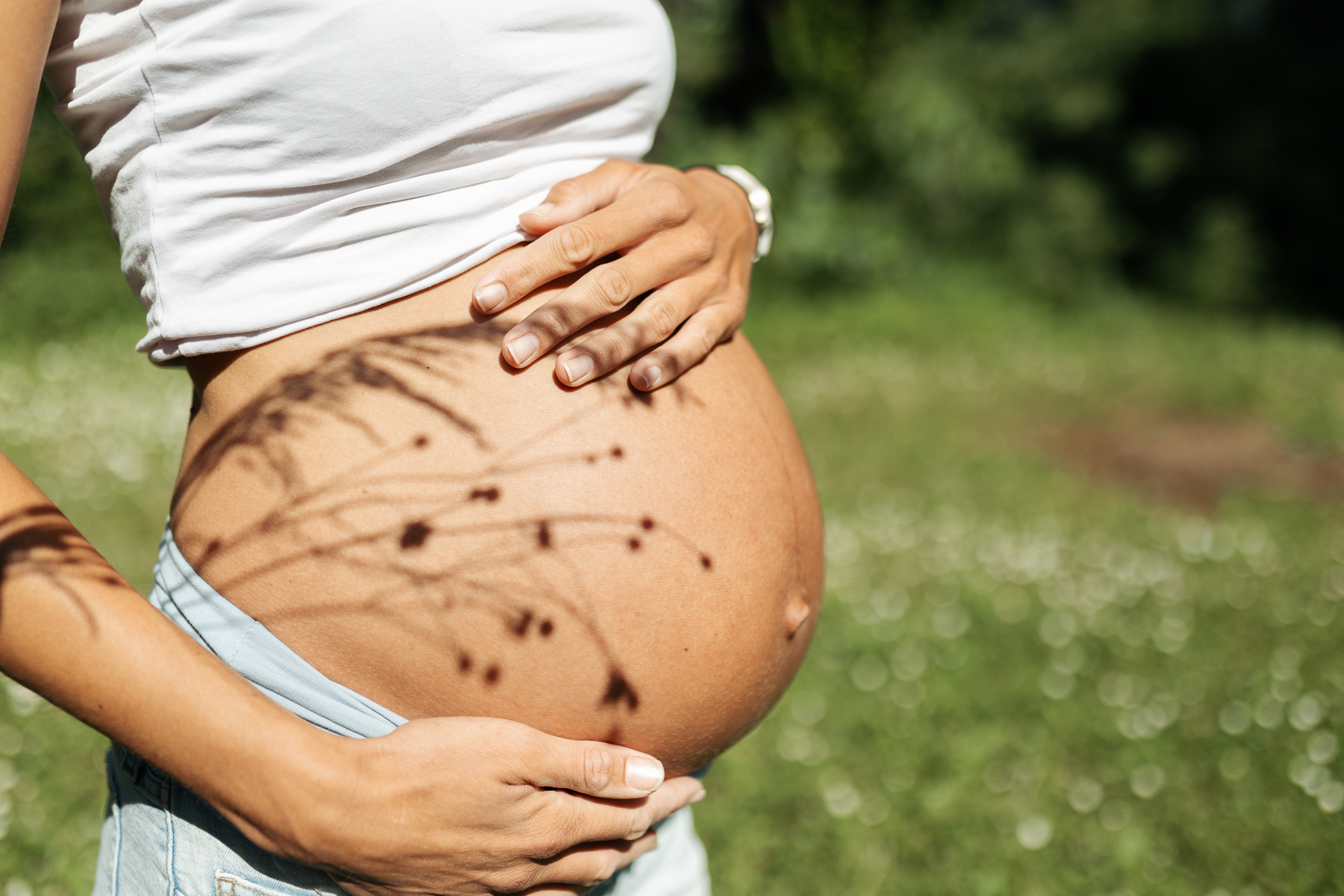 Nahaufnahme eines Babybauches im eigenen Garten im Kaiserstuhl. Auf dem Bauch sind Schatten von Gräsern zu sehen.
