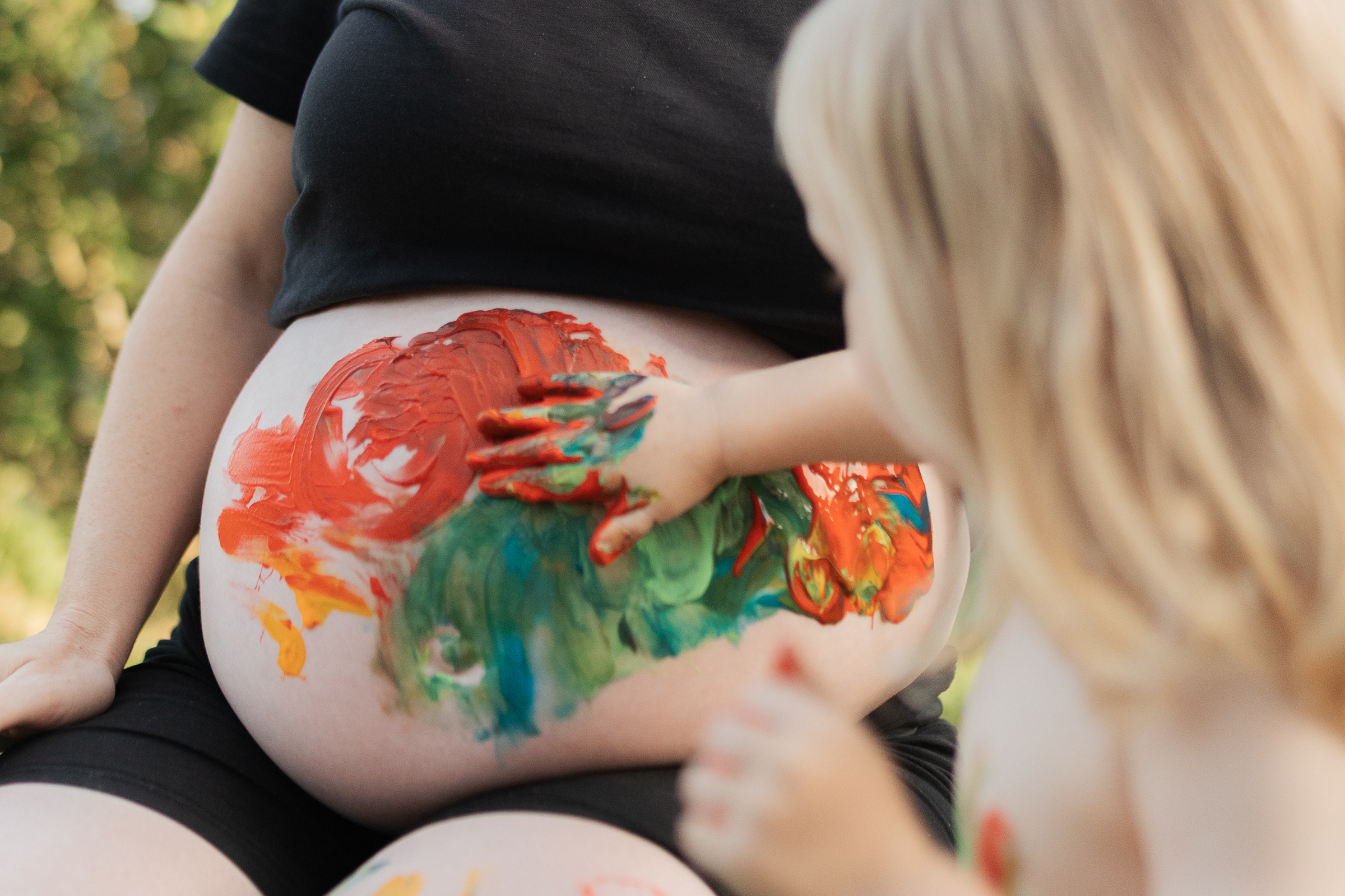 Schwangere Frau im Schwarzwald sitzt mit ihrer Tochter im Garten und bemalen den Babybauch mit Fingermalfarben.