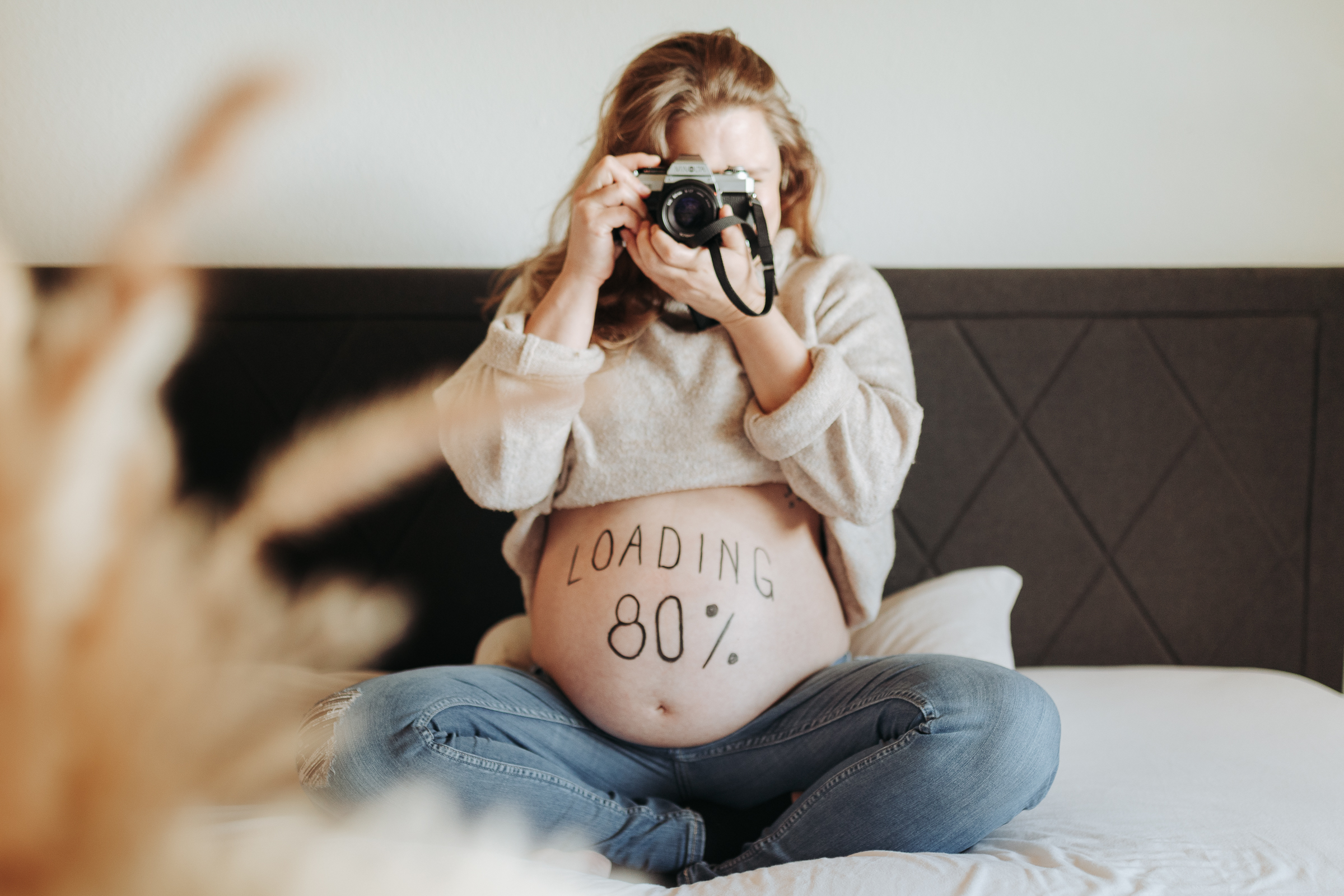 Werdende Mama mit Kamera auf dem Bett. Auf dem Bauch steht "Loading 80%"