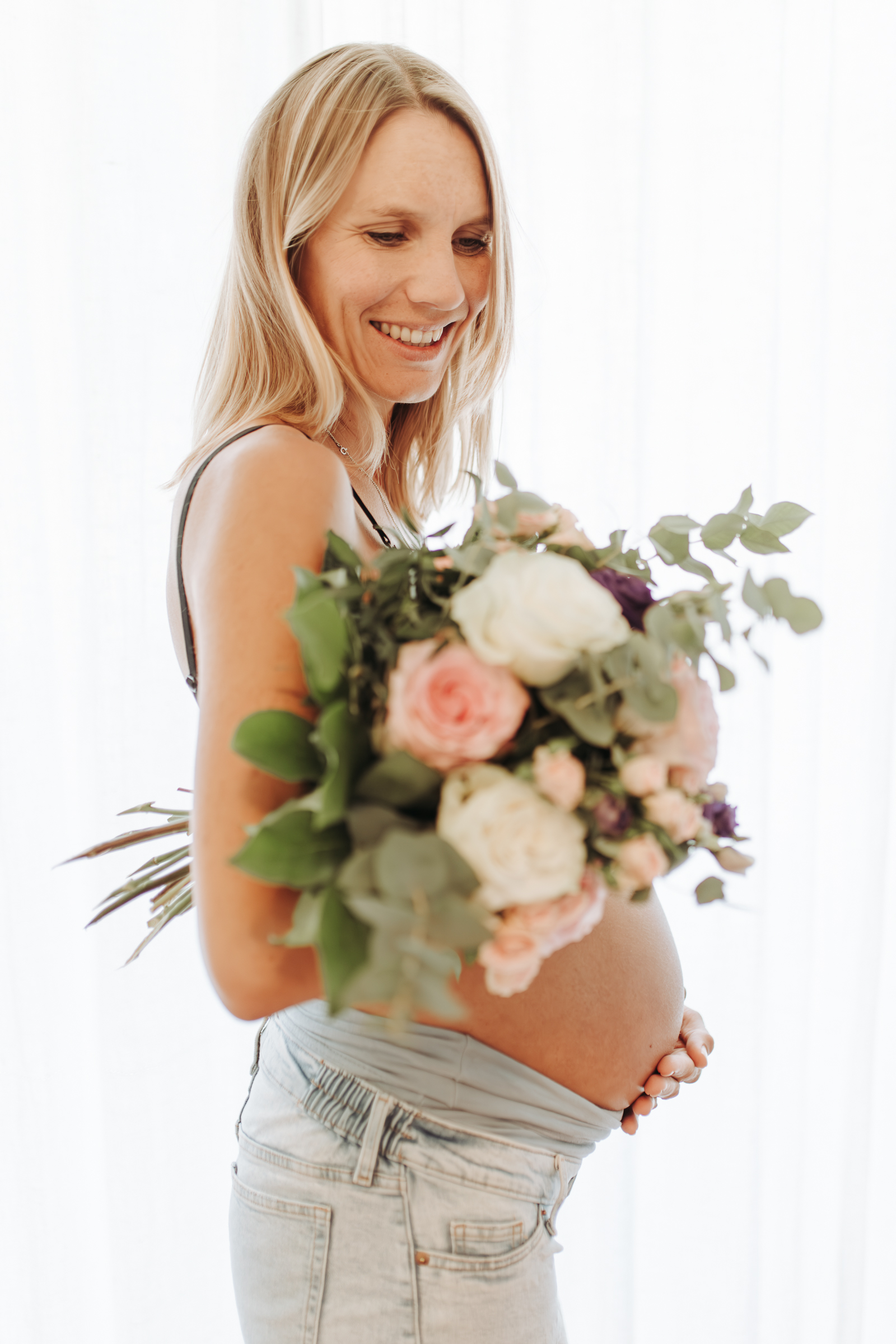 Werdende Mama aus dem Schwarzwald steht vor einem Fenster mit einem großen Blumenstrauß und hält ihren Babybauch.